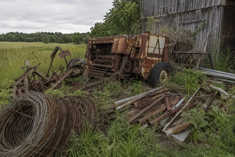 Old farm scrap metal, a rusted hay baler, implements, and fence wire piled near a weathered barn in a Lauderdale County field