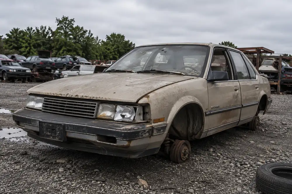 An old rusted junk car on a gravel lot waiting to be scrapped, bought as scrap weight against the current steel market